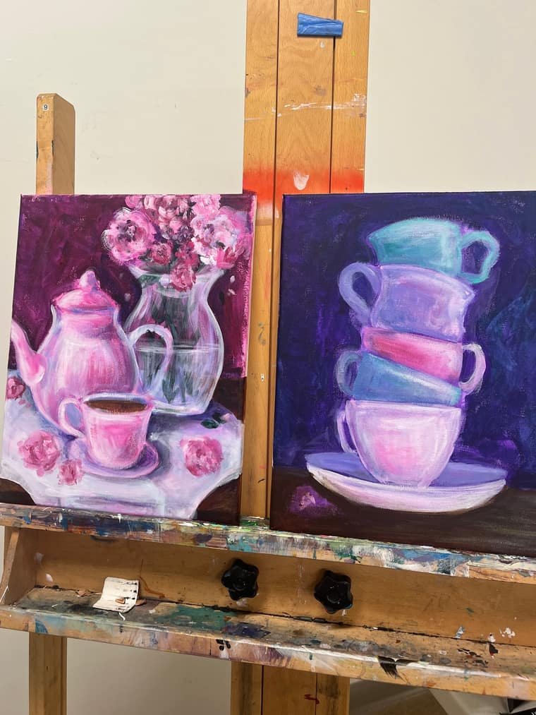 Two still life paintings on an easel: a pink teapot with roses and a stack of pastel teacups