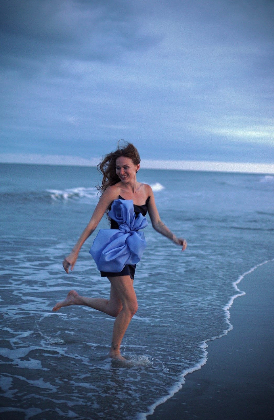 Model in a periwinkle bow dress running through the surf at dusk, from the Neverland collection