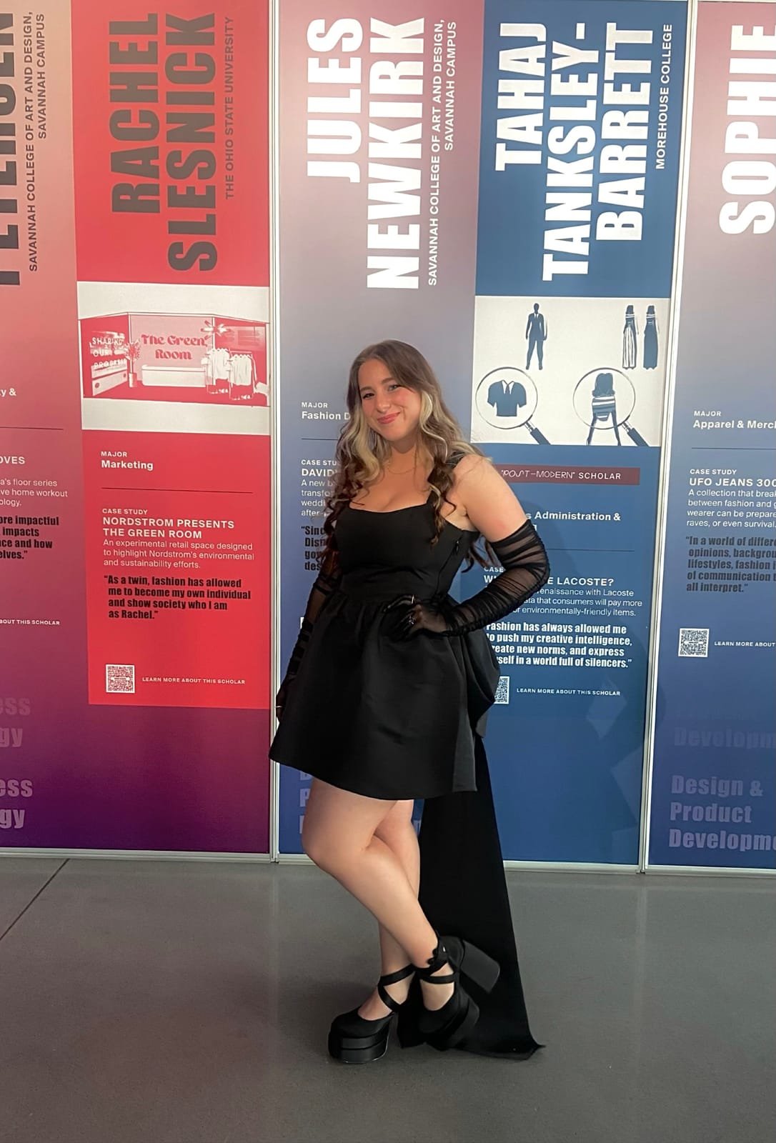 Jules in a black mini dress and long gloves in front of the Fashion Scholarship Fund scholar banners, her own behind her