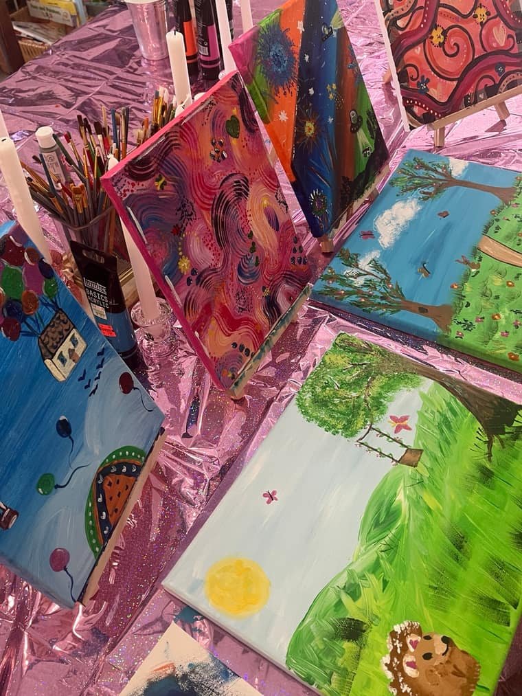 Six student canvases drying on a pink tablecloth after a painting class